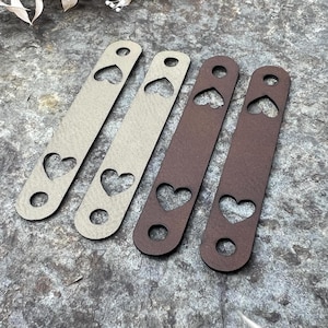 Puede incluir: Cuatro correas de cuero con recortes de corazón. Dos son de color beige claro y dos son de color marrón oscuro. Cada correa tiene un agujero en la parte superior e inferior. Las correas están dispuestas sobre una superficie gris texturizada.