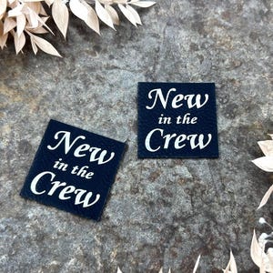 Könnte beinhalten: Zwei marineblaue quadratische Etiketten mit dem Text "New in the Crew" in weißer Schrift. Die Etiketten befinden sich auf einer strukturierten grauen Oberfläche, umgeben von getrocknetem beigem Laub.