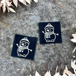 Könnte beinhalten: Zwei quadratische, marineblaue Stoffaufnäher mit einem weißen Schneemann-Design. Der Schneemann hat einen Hut, einen Schal und Knöpfe. Die Aufnäher liegen auf einer strukturierten, grauen Oberfläche, umgeben von getrocknetem, beigefarbenem Laub.