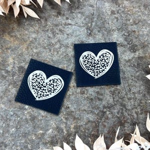 Könnte beinhalten: Zwei quadratische, marineblaue Aufnäher mit einem weißen Herzdesign, das mit einem Leopardenmuster gefüllt ist. Sie liegen auf einer strukturierten, grauen Oberfläche, mit getrockneten Blumendekorationen.