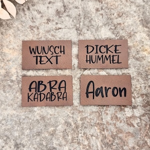 Könnte beinhalten: Vier kleine, rechteckige braune Lederetiketten mit schwarzem Text. Die Etiketten lauten "Wunsch Text", "Dicke Hummel", "Abra Kadabra" und "Aaron". Die Etiketten sind auf einer strukturierten, neutralfarbenen Oberfläche angeordnet.