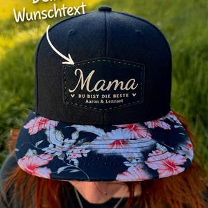 Könnte beinhalten: Schwarze Baseballkappe mit floral gemustertem Schirm. Auf der Kappe steht das Wort "Mama" in weißer Schrift auf einem rechteckigen Aufnäher. Unter dem Wort "Mama" steht der Text "DU BIST DIE BESTE" und die Namen "Aaron & Lennart".