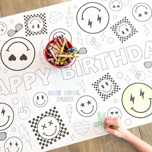 May include: A white coloring table cloth with the words "Happy Birthday" and "One Cool Dude" printed on it. The table cloth is decorated with various black and white smiley faces, hearts, stars, and lightning bolts. A hand is coloring in a peace sign with a green crayon.