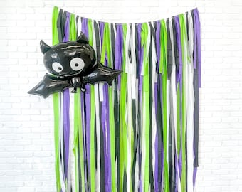May include: A black bat balloon with white eyes and a purple, green, black, and white streamer backdrop. The backdrop is made of thin strips of tissue paper.