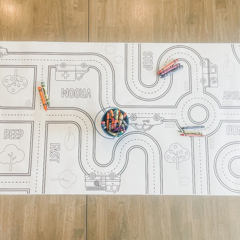 May include: A white colouring mat with black outlines of roads, cars, and trees. The mat has words like "Stop", "Vroom", "Beep", "Fast", "Honk", and "Hooray" printed on it. There are also crayons in a blue bucket and on the mat.
