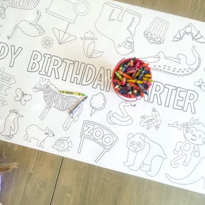 May include: A white coloring table cloth with a zoo theme. The table cloth features a variety of animals including a zebra, a monkey, a panda, a snake, a frog, and an alligator. The table cloth also includes the words "Happy Birthday Carter" and "Zoo". A red bucket filled with crayons is on the table cloth.