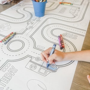 May include: A white colouring mat with a road and car theme. The mat has a black and white outline of a road with stop signs, traffic lights, and cars. The text "Birthday" is written on the mat. A child's hand is colouring the mat with blue crayons.