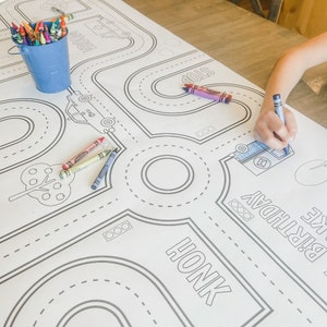 May include: A white tablecloth with a black and white road design for colouring. The design includes a tree, a car, a truck, and the words "Honk" and "Stop". There are also crayons in a blue bucket.