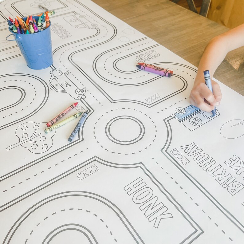 May include: A white tablecloth with a black and white road design for colouring. The design includes a tree, a car, a truck, and the words "Honk" and "Stop". There are also crayons in a blue bucket.