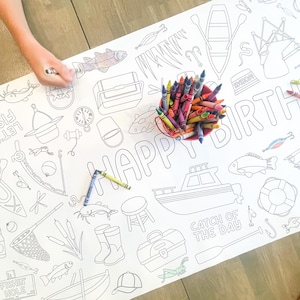May include: A white paper coloring sheet with a fishing theme, featuring outlines of fish, boats, and fishing gear. The words "HAPPY BIRTH" are printed in large letters, with a red container of crayons nearby. A hand is coloring the sheet.
