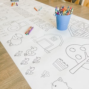 May include: A white colouring tablecloth with a farm theme. The tablecloth features a tractor, a barn, a sheep, ducks, an apple tree, a flower, and a cat. The text "Happy Birthday Kyler" is printed on the tablecloth. A bucket of crayons is on the table.