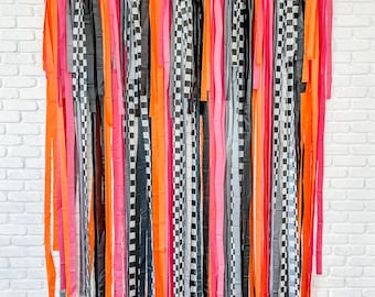 May include: A black and white checkered, pink, orange, and gray streamer backdrop. The streamers are made of tissue paper and are hanging from a white wall.