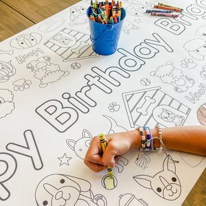 Pu&ograve; includere: Un foglio da colorare bianco con un tema di compleanno di cane. Il foglio presenta una variet&agrave; di illustrazioni di cani, tra cui una cuccia, ossa e impronte di zampe. Il testo "Buon compleanno" &egrave; scritto in cima al foglio. Una mano di bambino sta colorando il foglio con un pastello verde.
