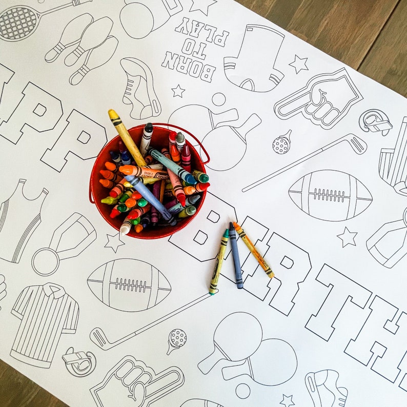 A white table cover with a black and white sports-themed design. The words "Born to Play" are printed on the cover. A red bucket filled with crayons is on the table cover.