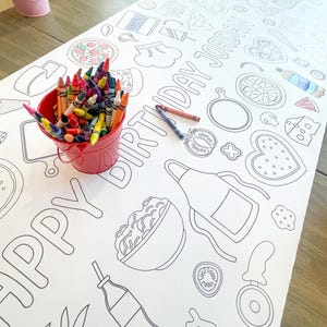 A large white paper tablecloth with black line drawings of food items and the text "HAPPY BIRTHDAY JORDAN". A red bucket filled with colorful crayons sits on the table. Two crayons are next to the bucket.