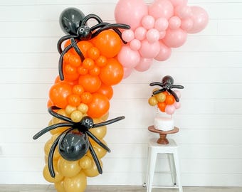 May include: A Halloween balloon arch with orange, gold, and black balloons. The arch is decorated with black spider balloons. A smaller balloon arch with pink, orange, and gold balloons is on a white cake stand.