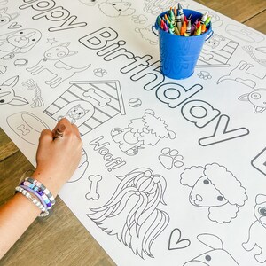Pu&ograve; includere: Una pagina da colorare bianca con le parole "Buon compleanno" in lettere nere delineate. La pagina presenta varie illustrazioni a tema cani, tra cui una cuccia per cani, ossa, impronte di zampe e diverse razze di cani. Un secchio blu pieno di matite colorate si trova nell'angolo in alto a destra della pagina.