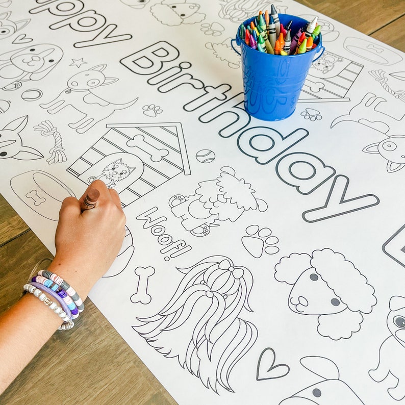 Pu&ograve; includere: Una pagina da colorare bianca con le parole "Buon compleanno" in lettere nere delineate. La pagina presenta varie illustrazioni a tema cani, tra cui una cuccia per cani, ossa, impronte di zampe e diverse razze di cani. Un secchio blu pieno di matite colorate si trova nell'angolo in alto a destra della pagina.