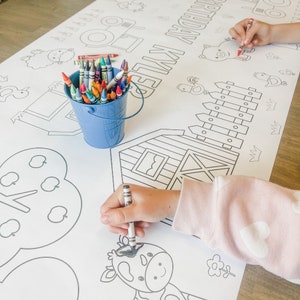 May include: A white colouring tablecloth with a farm theme. The tablecloth features a variety of farm animals, including a pig, a cow, a chicken, and a tractor. The text "Happy Birthday" is printed on the tablecloth. A bucket of crayons is on the table, and a child is colouring on the tablecloth.