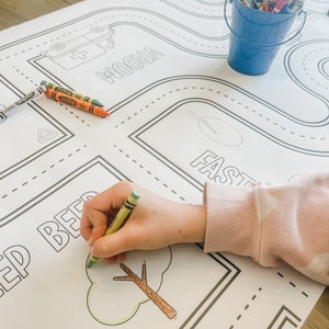 May include: A child's hand colours a tree on a large, white, printed play mat. The mat features a road with the word "VROOM" and a tree with the word "FAST".