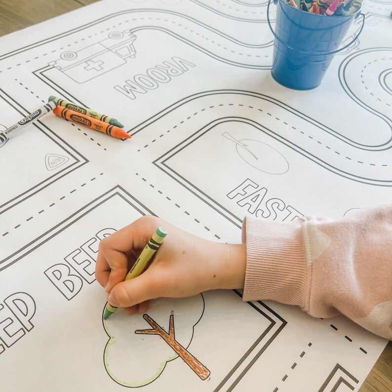 May include: A child's hand colours a tree on a large, white, printed play mat. The mat features a road with the word "VROOM" and a tree with the word "FAST".