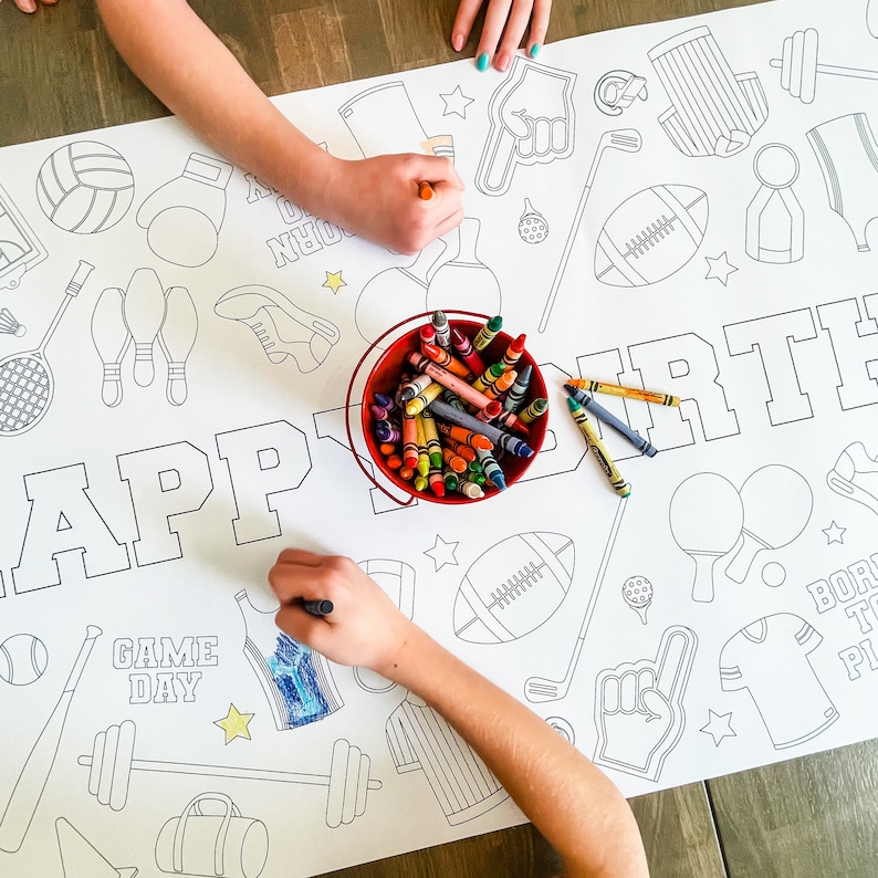 A white coloring page with the words "Happy Birthday" and "Game Day" printed on it. The page is decorated with various sports-themed illustrations, including a football, baseball bat, basketball, bowling pins, and a golf club. A red bucket filled with crayons is in the center of the page.