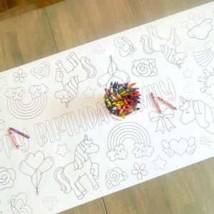 May include: A white table runner with a unicorn theme. The runner features a variety of images including unicorns, rainbows, stars, hearts, flowers, and a bow. The text "Happy Birthday" is written across the runner. A bowl of crayons is on top of the runner.