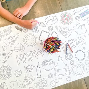 A white paper tablecloth with a pizza-themed design, featuring pizza slices, ingredients, and the words "HAPPY BIRTHDAY" and "PARTY TIME". A red container of crayons and two crayons are next to the tablecloth. A person is coloring the tablecloth.