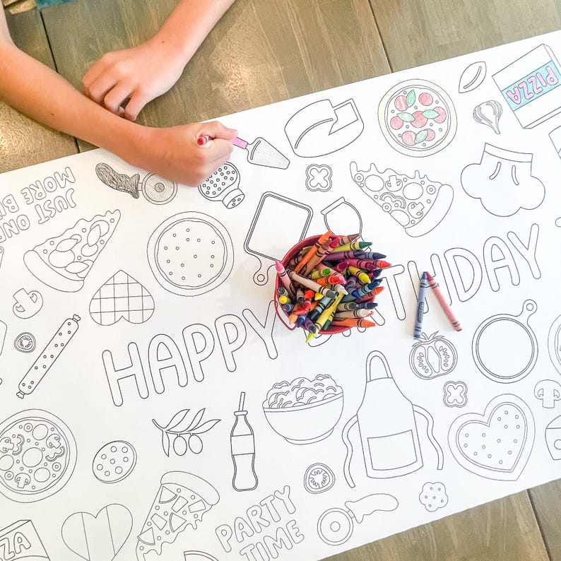 A white paper tablecloth with a pizza-themed design, featuring pizza slices, ingredients, and the words "HAPPY BIRTHDAY" and "PARTY TIME". A red container of crayons and two crayons are next to the tablecloth. A person is coloring the tablecloth.