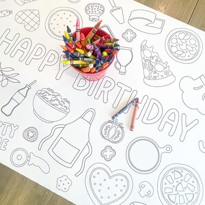 A white paper tablecloth with a black outline design of pizza-themed illustrations and the words "HAPPY BIRTHDAY". A red bucket filled with colorful crayons sits on the tablecloth, with two crayons lying on the surface.