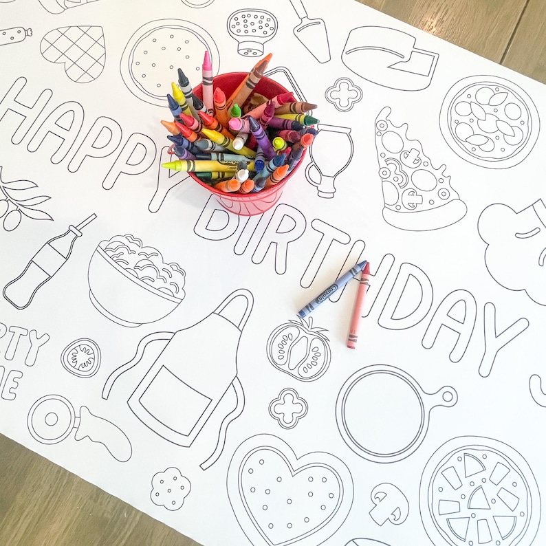 A white paper tablecloth with a black outline design of pizza-themed illustrations and the words "HAPPY BIRTHDAY". A red bucket filled with colorful crayons sits on the tablecloth, with two crayons lying on the surface.