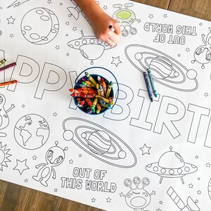 May include: A white coloring table cloth with an outer space theme. The table cloth features the words "Happy Birthday" and "Out of this World" with planets, stars, and aliens. A child's hand is coloring the table cloth with crayons.