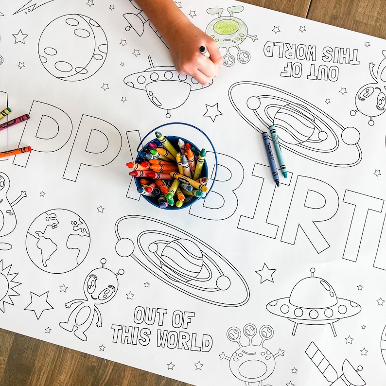 May include: A white coloring table cloth with an outer space theme. The table cloth features the words "Happy Birthday" and "Out of this World" with planets, stars, and aliens. A child's hand is coloring the table cloth with crayons.