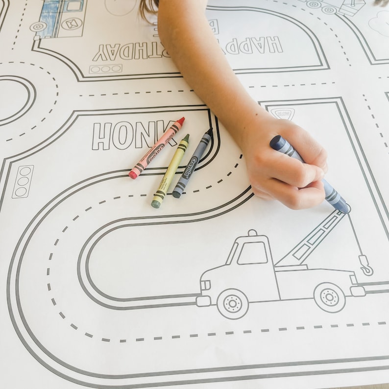 May include: A white colouring mat with a black and white road design. The mat features a tow truck, a bus, and the words "Happy Birthday" and "Honk". A child's hand is holding a blue crayon and colouring the tow truck.