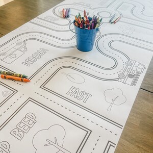 May include: A black and white colouring mat featuring a road with various words like "STOP", "VROOM", "FAST", and "BEEP". There are also images of trees, a fire engine, and an ambulance. A blue bucket with coloured crayons is in the top left corner of the mat.