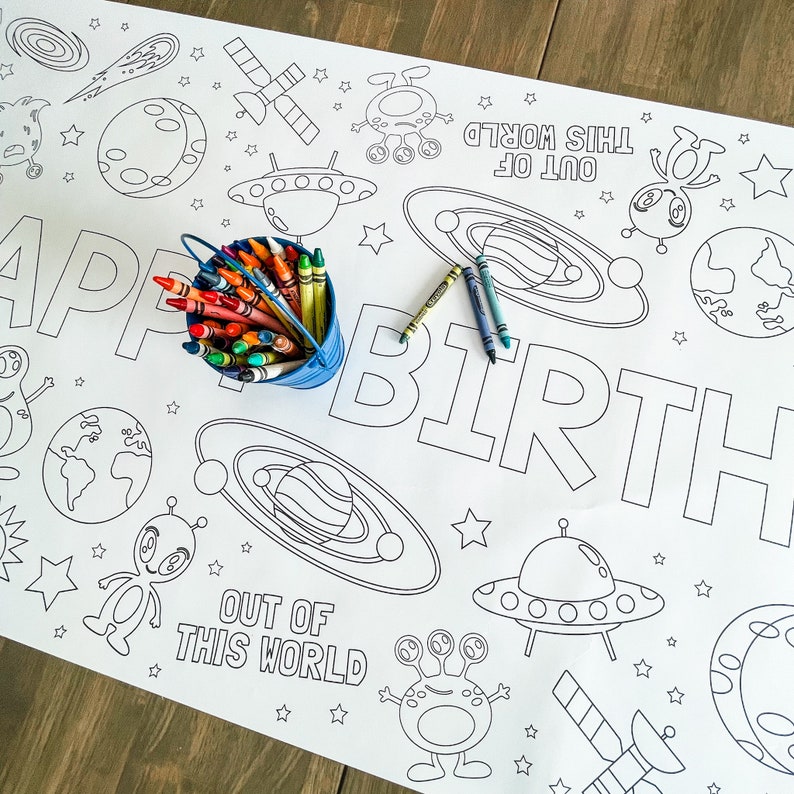May include: A black and white coloring page with an outer space theme. The page features the words "Happy Birthday" and "Out of this World" along with images of planets, stars, spaceships, and aliens. A box of crayons is sitting on the page.