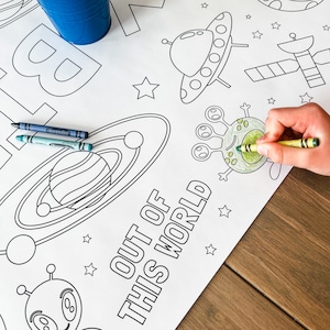 May include: A black and white coloring page with the text "Out of this world". The page features a spaceship, a planet, a satellite, and an alien. Crayola crayons are on the page.