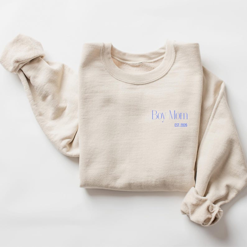 May include: A cream-colored sweatshirt with the text "Boy Mom" in blue script, with "EST. 2026" below. The sleeves are rolled up. The sweatshirt is made of a soft material.