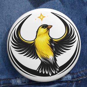 Pode incluir: Um botão redondo com um desenho estilizado de um pássaro amarelo. O pássaro tem asas pretas e amarelas, uma cabeça preta e um bico laranja. Uma estrela dourada está acima do pássaro. O botão está sobre um fundo de ganga azul.