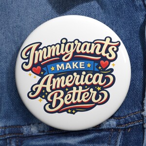 Könnte beinhalten: Ein weißer Button mit dem Text "Immigrants Make America Better" in einer stilisierten Schriftart. Der Text ist in Rot, Blau und Gelb gehalten, mit Herz- und Sternakzenten. Der Button befindet sich vor einem blauen Jeanshintergrund.
