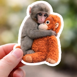 May include: A sticker featuring a gray monkey hugging an orange stuffed animal monkey. The sticker has a white border and is held by a person's hand. The background is blurred green.