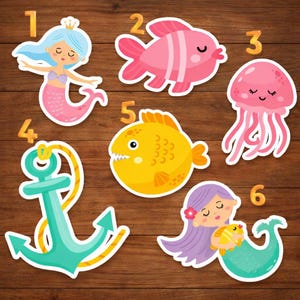 May include: A collection of colorful, cartoon-style stickers featuring marine life. Includes a mermaid with a crown, a pink fish, a jellyfish, a teal anchor, a yellow fish, and a mermaid holding a fish. The stickers are numbered 1 through 6.
