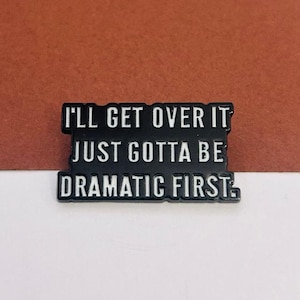 May include: A black enamel pin with the text "I'LL GET OVER IT JUST GOTTA BE DRAMATIC FIRST" in white lettering. The pin is rectangular with rounded corners and is set against a brown and white background.