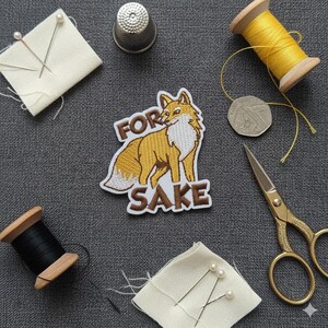 May include: Embroidered fox patch with the words "FOR SAKE". The image shows sewing items: yellow and black thread spools, a thimble, scissors, pins, and a coin, arranged on a grey surface. A craft-themed composition.