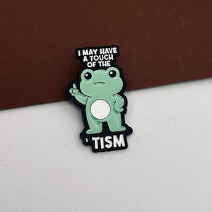 May include: A green frog-shaped badge with the text "I MAY HAVE A TOUCH OF THE 'TISM" in white. The frog is outlined in black and has a white belly. The frog is giving a thumbs up.