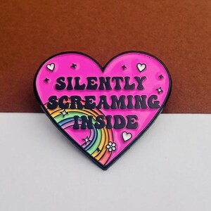 May include: A heart-shaped enamel pin with a black border and a bright pink background. The pin features the words "SILENTLY SCREAMING INSIDE" in black, along with a rainbow, stars, hearts, and a flower design.