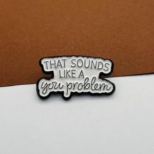 May include: A white enamel pin with the phrase "THAT SOUNDS LIKE A you problem" in black lettering. The pin has a black border. The pin is set against a brown background. A fun accessory.