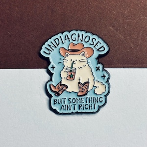 May include: A light blue enamel pin featuring a fluffy cream-coloured cat wearing a brown cowboy hat and matching boots. The cat holds a drink with a green straw. The text reads UNDIAGNOSED BUT SOMETHING AIN'T RIGHT in black capital letters.