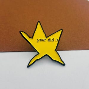 May include: A yellow star-shaped enamel pin with a black outline. The pin features the phrase "your did it" in a black, sans-serif font. The pin is set against a brown and white background.
