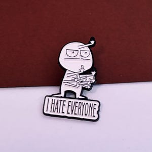 May include: A white and black enamel pin depicting a cartoon figure with a raised middle finger. The figure has a simple design and the pin includes the text "I HATE EVERYONE". A humorous accessory.